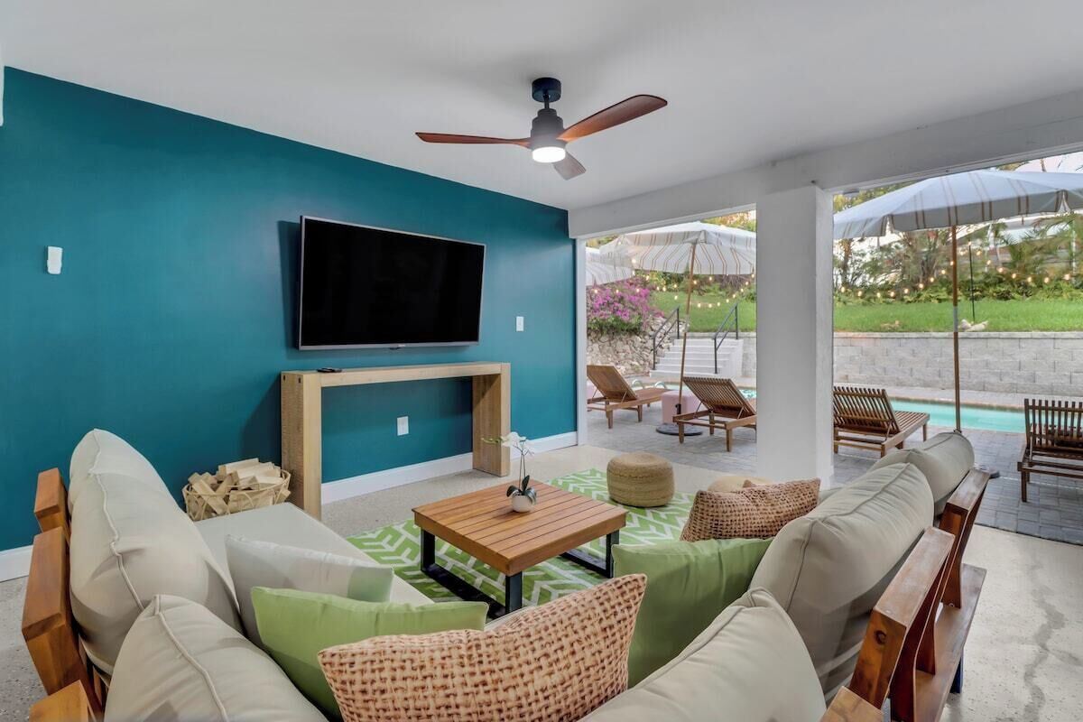 A modern living room with a flat-screen TV, a ceiling fan, and a view of an outdoor patio with lounge chairs and a pool.