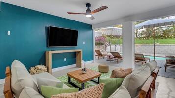 A modern living room with a flat-screen TV, a ceiling fan, and a view of an outdoor patio with lounge chairs and a pool.
