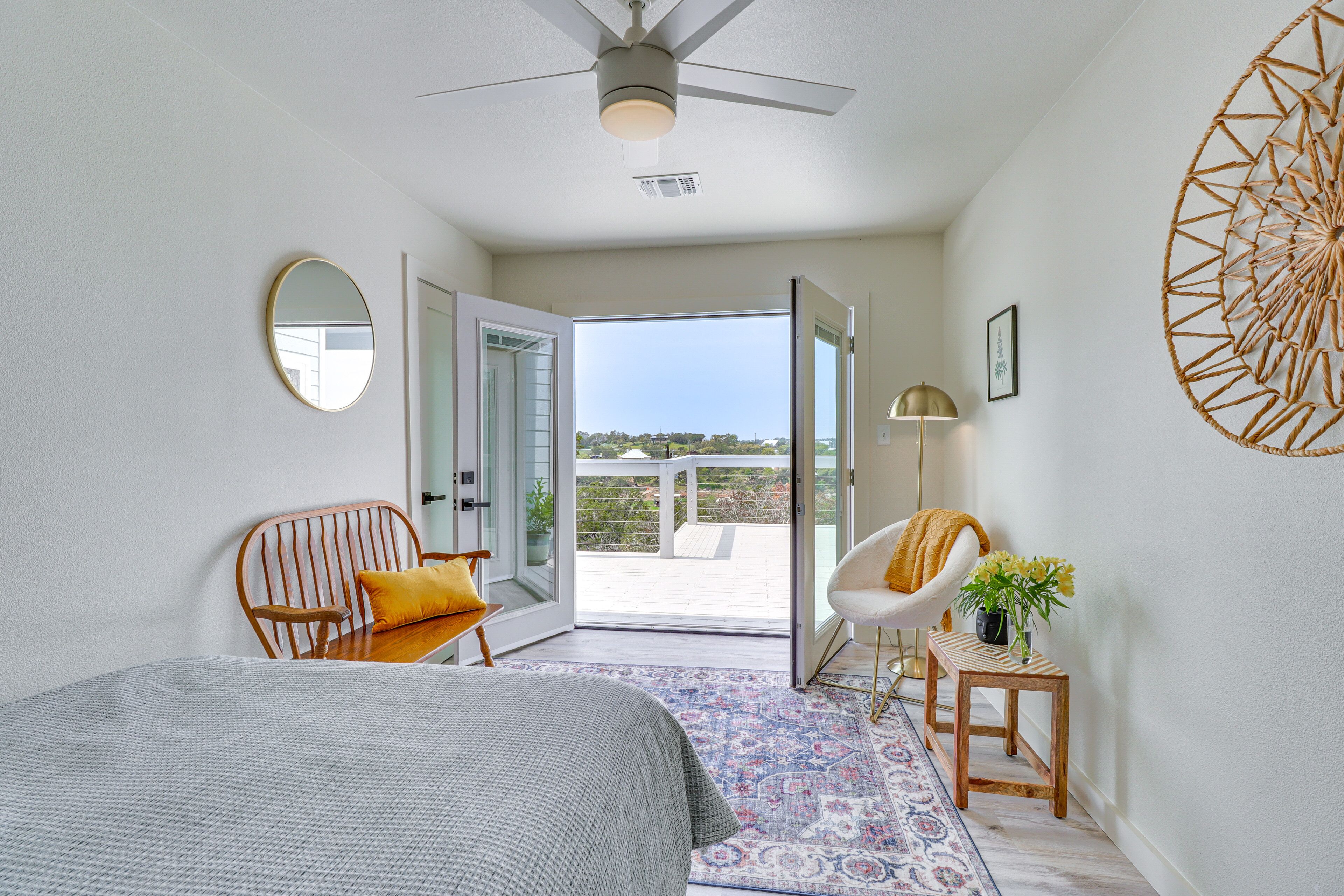 A bedroom with a bed, a chair, a small table with a plant, a mirror, and a view of the outdoors.