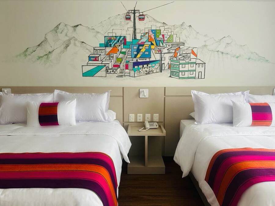Quarto de hotel com duas camas, cada uma com roupa de cama branca e cobertores listrados coloridos. Pequena mesa de cabeceira com telefone e interruptor de luz na parede. Acima das camas, um mural colorido retrata uma paisagem de montanhas com edifícios e um teleférico.