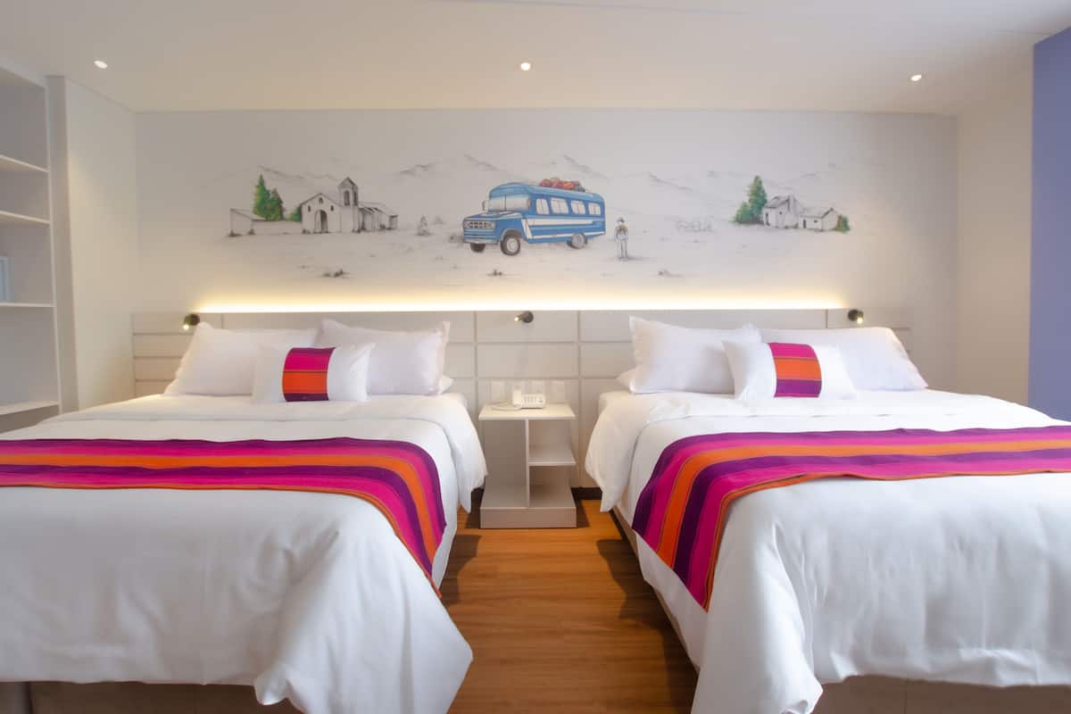 A hotel room with two beds, a nightstand, a wall-mounted shelf, and a mural depicting a bus and mountains.