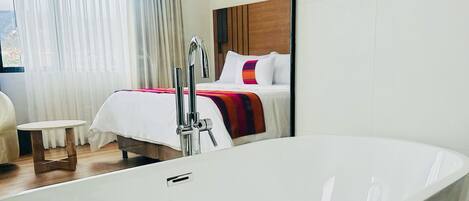 Habitación de hotel moderna con una bañera independiente grande, cabecero de madera y un amplio ventanal con cortinas de gasa.