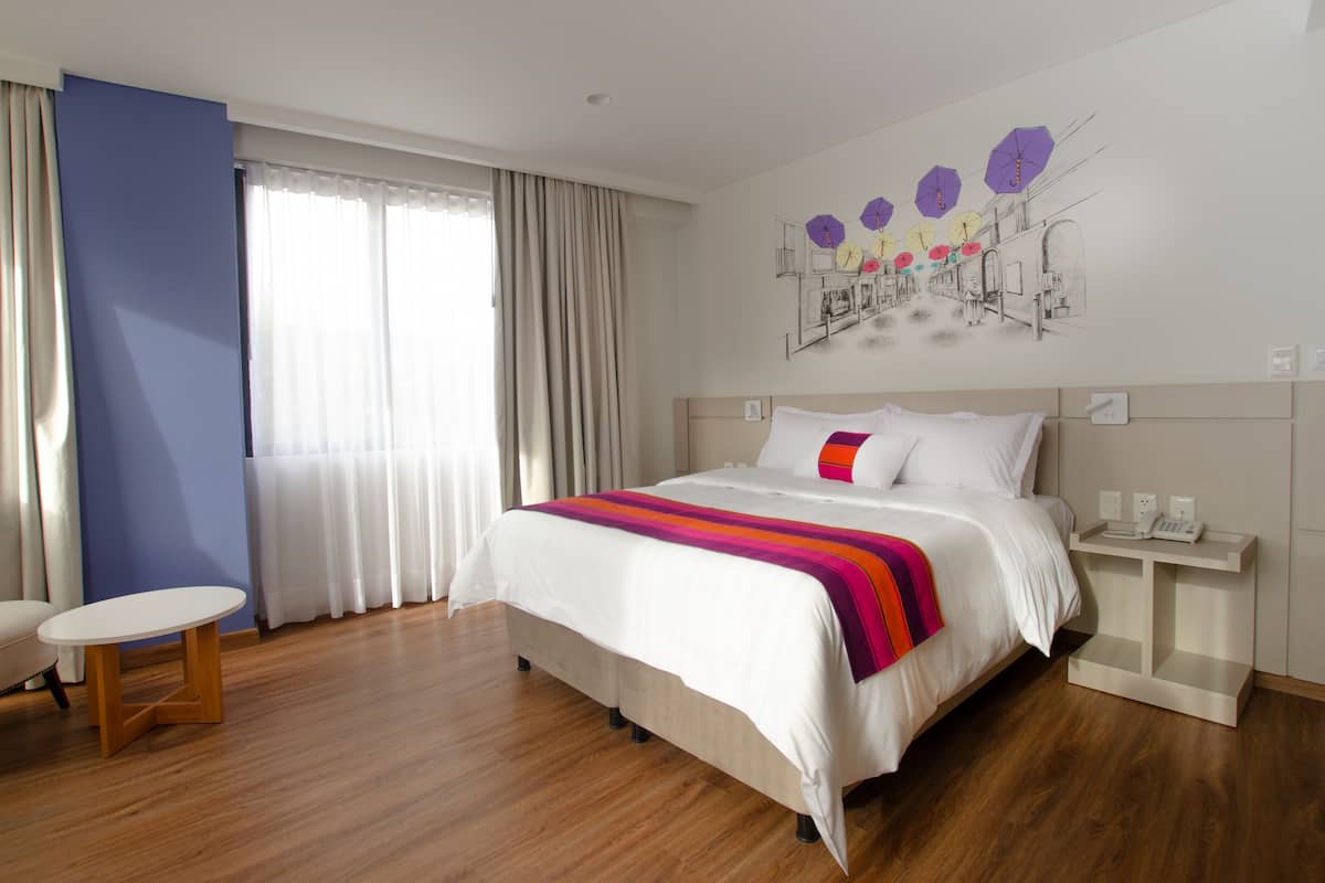 A modern hotel room with a large bed, a bedside table, a small round table, and a large window with curtains.