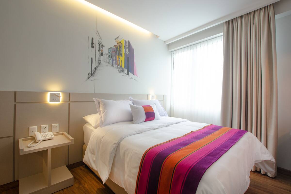 A hotel room with a neatly made bed, a bedside table with a phone, and a wall-mounted light.