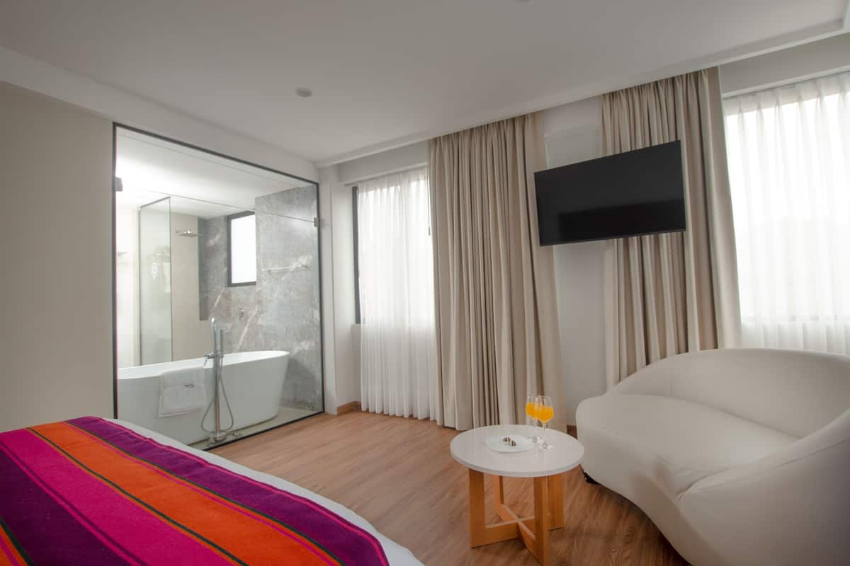 A modern hotel room with a large bed, a white armchair, a small round table with a plate and glasses, a flat-screen TV mounted on the wall, and a glass-enclosed shower area.