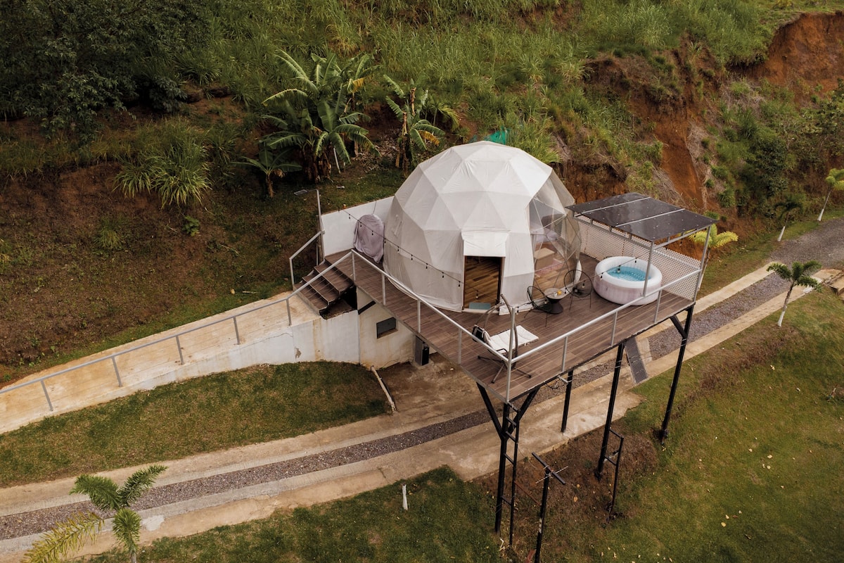 Une structure en forme de dôme géodésique dotée d’un bain à remous et d’un espace de détente extérieur, entourée d’une terrasse en bois et d’un sentier.