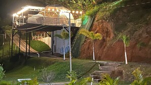 A multi-level outdoor structure with a covered seating area, surrounded by greenery and a hillside.