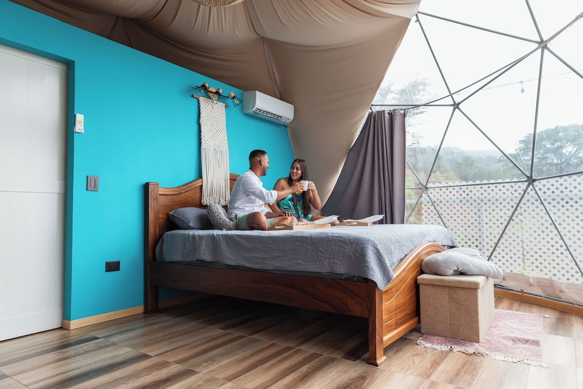 Une chambre avec un lit en bois, un mur de couleur turquoise et une vue à travers une structure de fenêtre aux motifs géométriques.