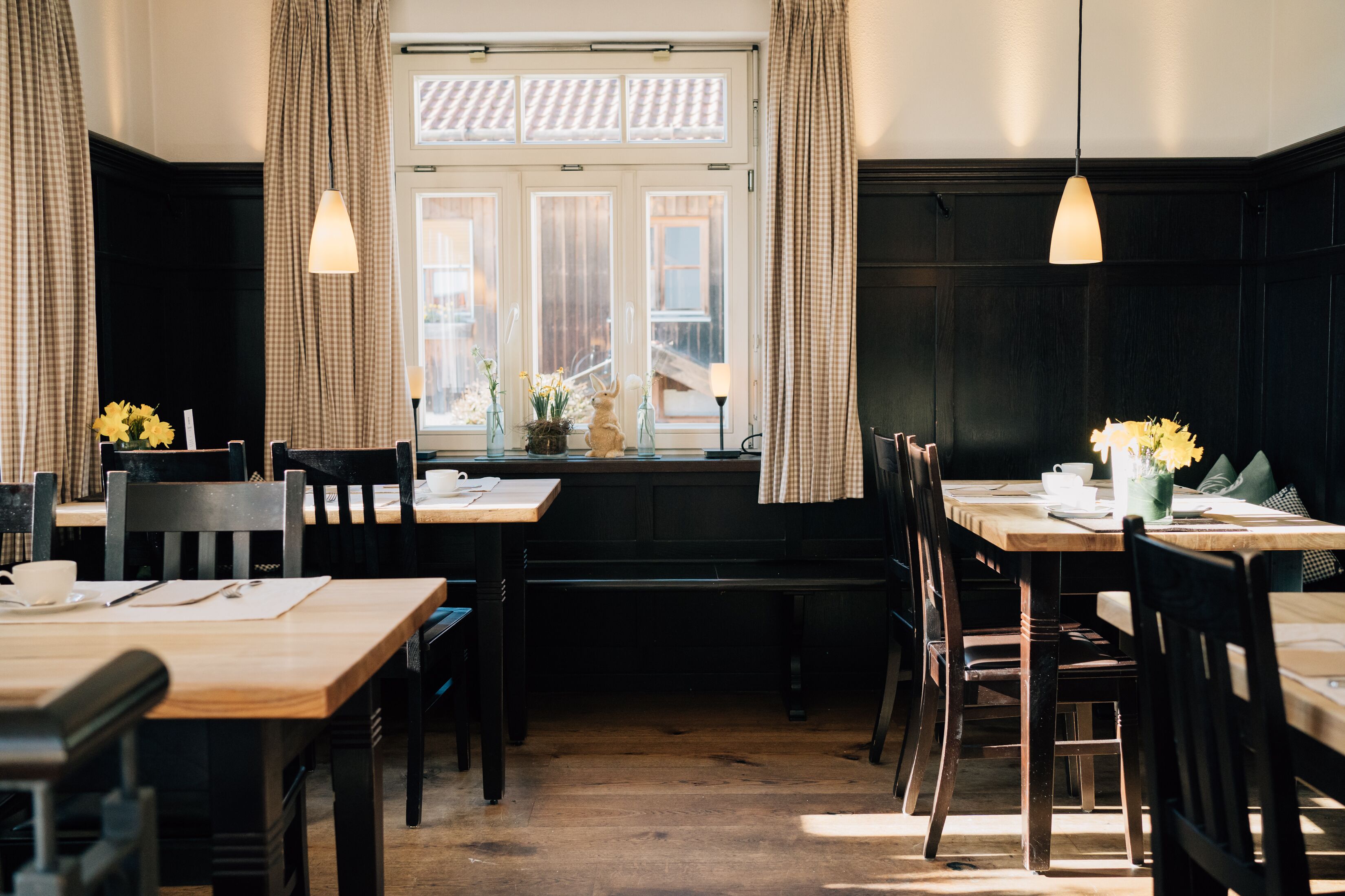 A cozy dining area with wooden tables and chairs, a window with curtains, and pendant lights.