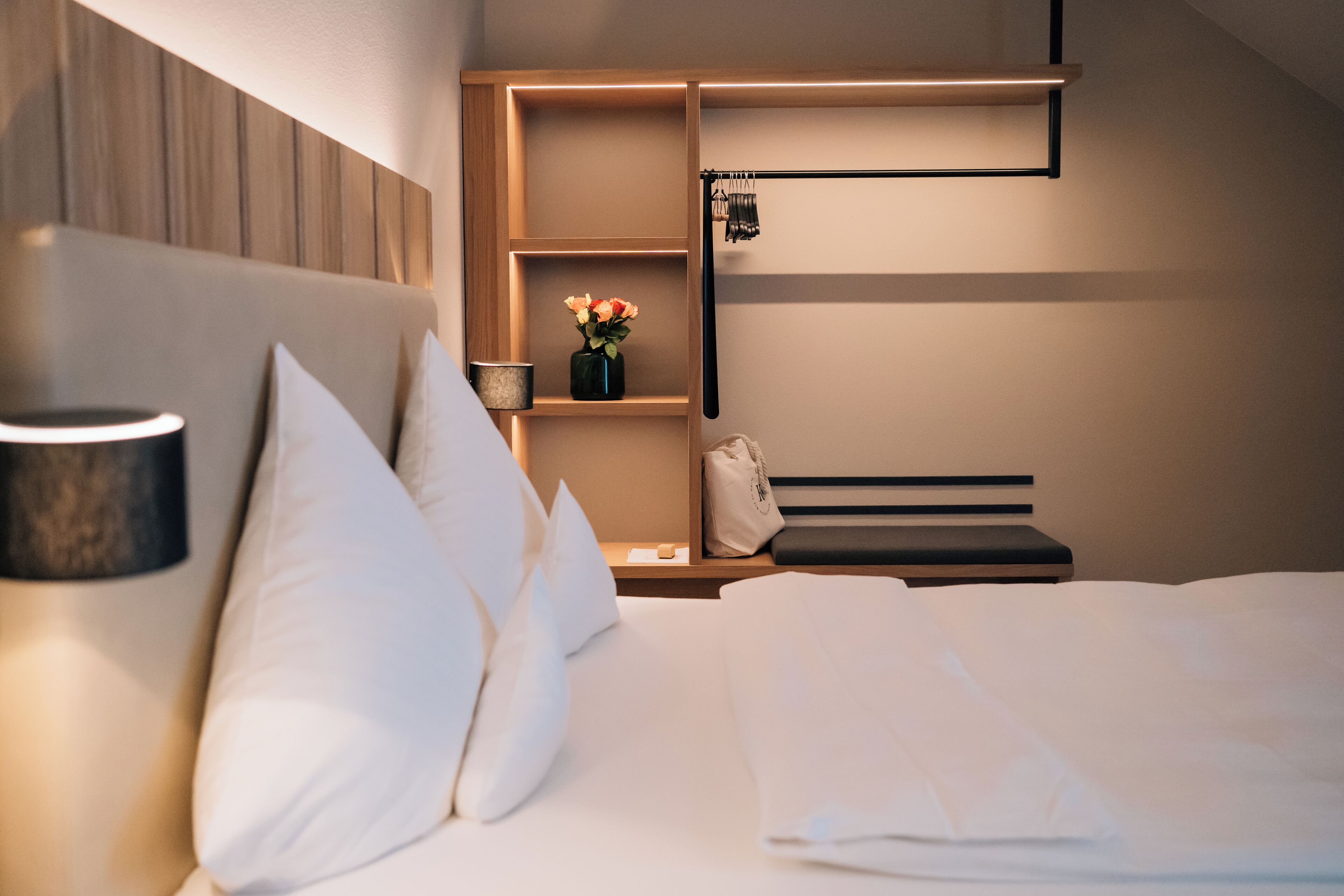 A modern hotel room with a neatly made bed, a bedside table with a lamp, a shelf with a vase of flowers, and a hanging coat rack.