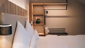 A modern hotel room with a neatly made bed, a bedside table with a lamp, a shelf with a vase of flowers, and a hanging coat rack.
