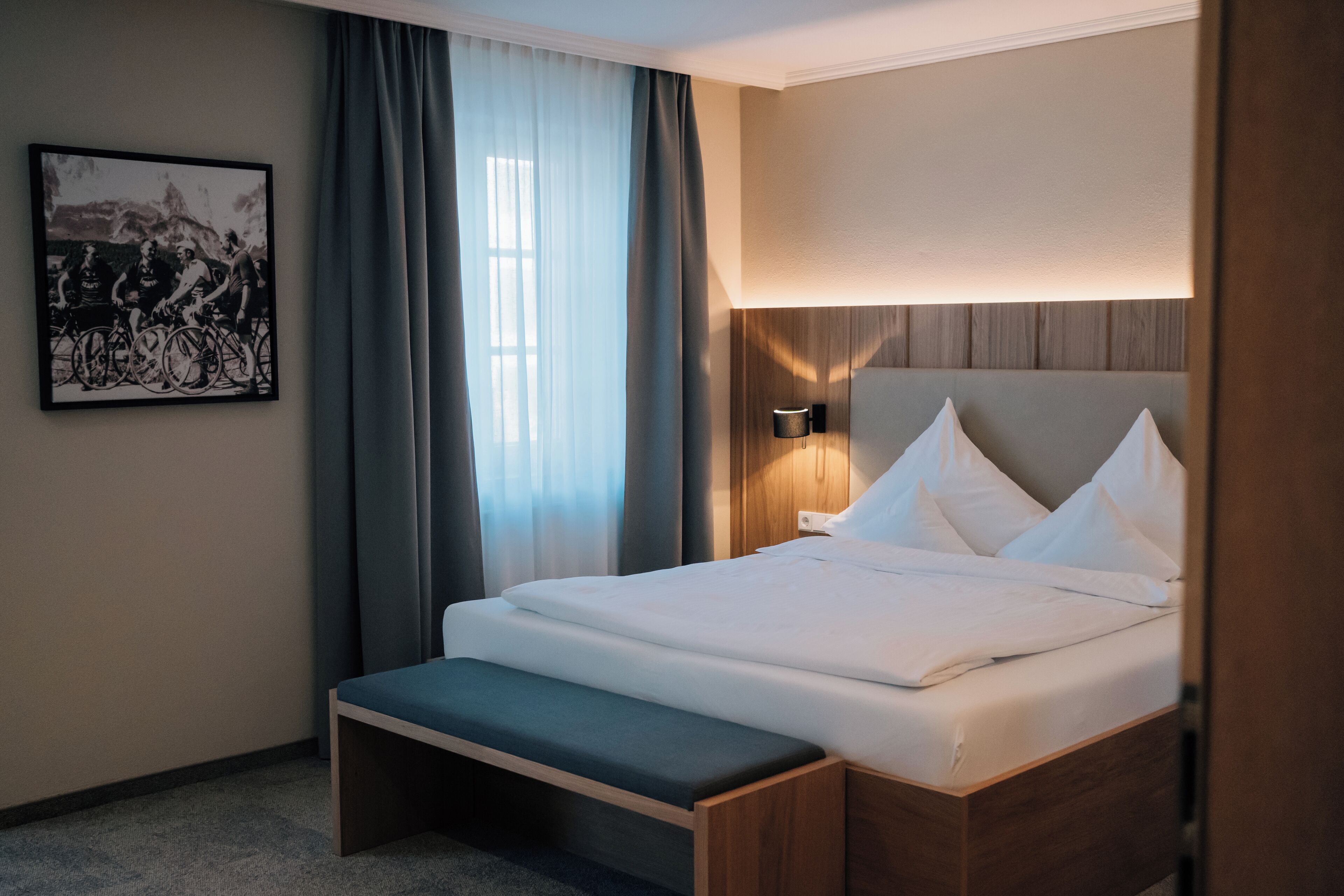 A modern hotel room with a wooden bed, a bench, a wall-mounted lamp, and a framed picture.