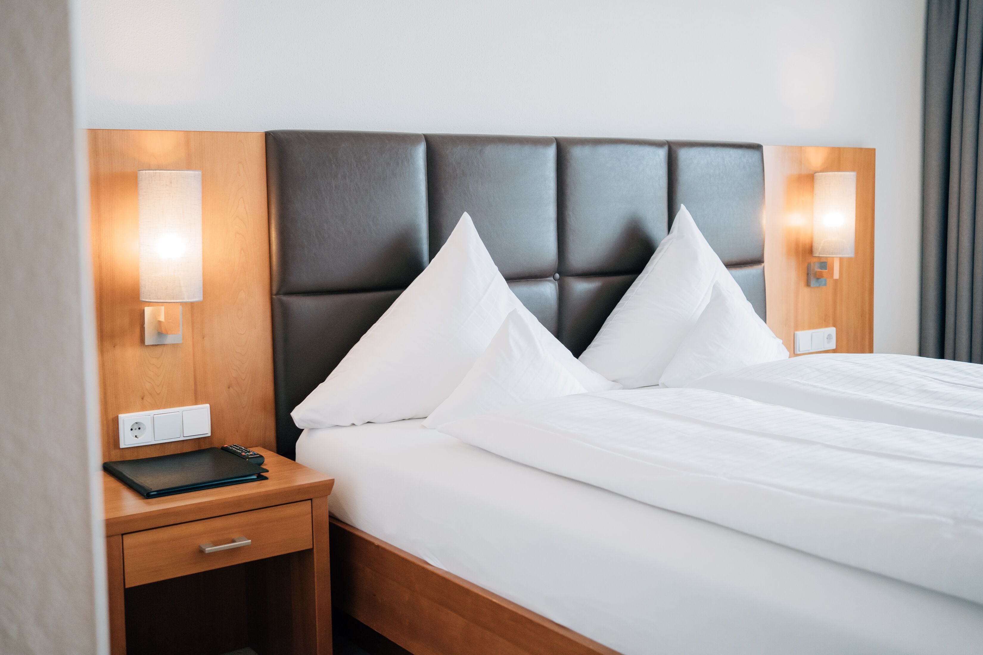 Cama bem arrumada com lençóis brancos e cabeceira escura. Há duas mesas de cabeceira, cada uma com uma luminária e uma tomada.