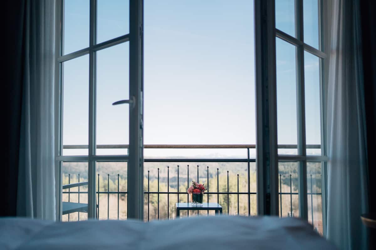 A room with a bed, a window with a view of a mountainous landscape, and a small table with a vase of flowers.