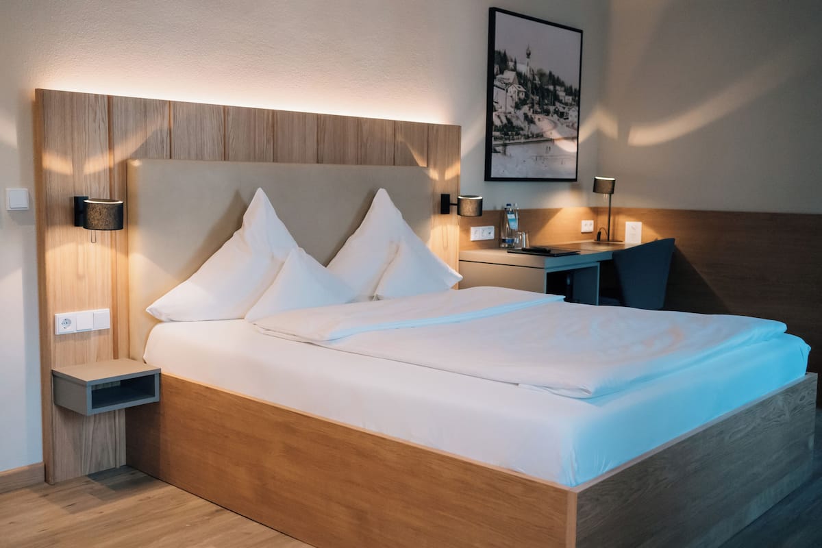 A modern hotel room with a wooden headboard, a neatly made bed with white linens, a small bedside table with a lamp, and a framed picture on the wall.