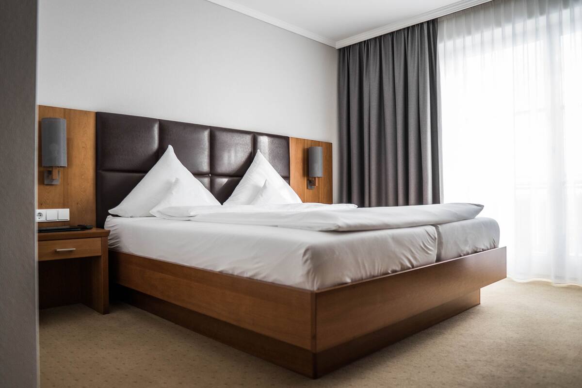 A modern hotel room with a large bed, wooden headboard, two bedside tables, and a view of the outdoors through sheer curtains.