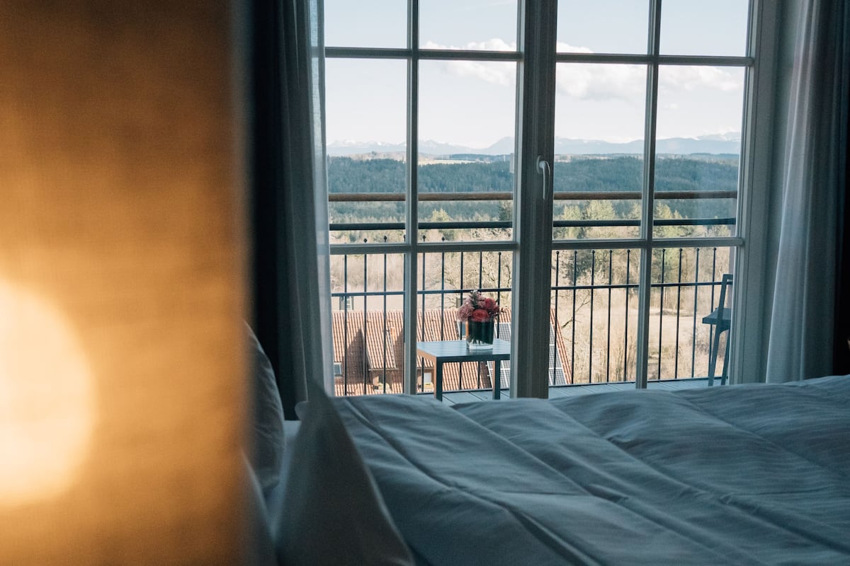 A bedroom with a large window offering a view of a fenced outdoor area and distant mountains.