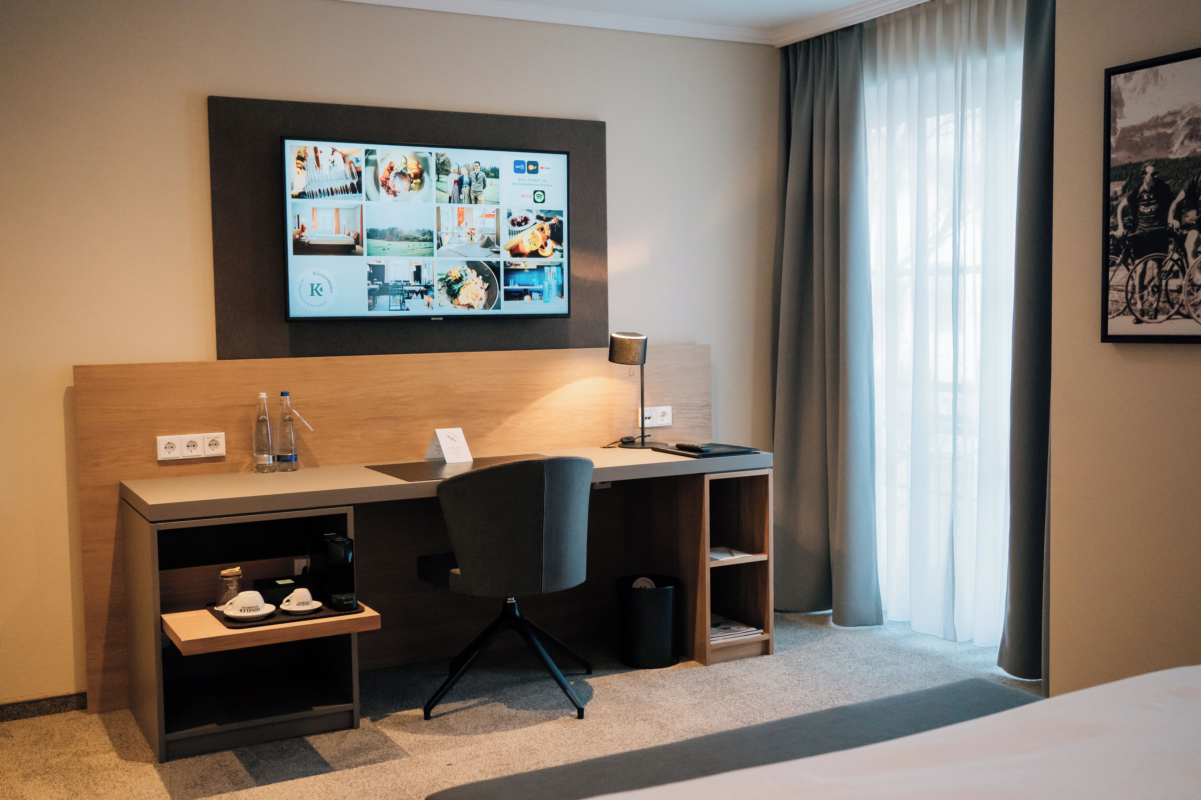 A hotel room with a wooden desk, a chair, a flat-screen TV mounted on the wall, a bed, and a window with curtains.