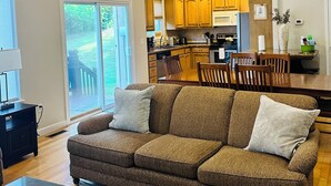 Een ruime woonkamer met een grote bruine bank, een eettafel met stoelen, en een keuken met houten kasten en uitzicht op de omgeving.