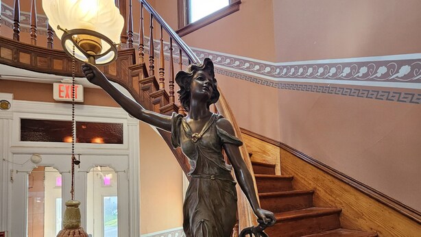 Une statue d’une femme tenant une lanterne sur un escalier doté d’une rampe en bois et d’une porte blanche avec des panneaux de verre.
