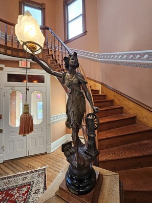 A statue of a woman holding a lantern on a staircase with a wooden railing and a white door with glass panels. - Starrett House  (Port Townsend)