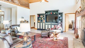 Ein geräumiges Wohnzimmer mit hoher Decke, Kamin, großem Fernseher und einer gemütlichen Sitzecke mit Ledersofas und einem gemusterten Teppich.