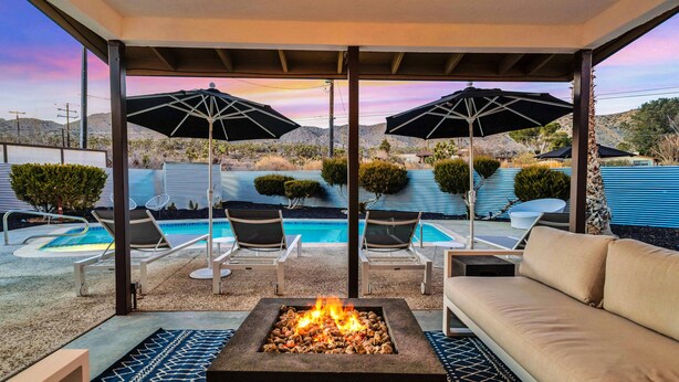 Uma área de estar coberta ao ar livre com lareira, cadeiras de descanso e guarda-sóis, com vista para uma piscina e paisagem desértica.