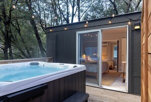 A hot tub outside a wooden cabin with a glass door leading to a bedroom with a bed and a nightstand. - Modern Cabin (Redwood) (Dunsmuir)