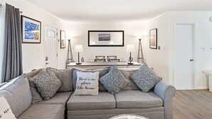 A living room with a grey sofa, two lamps, framed pictures on the wall, and a door.