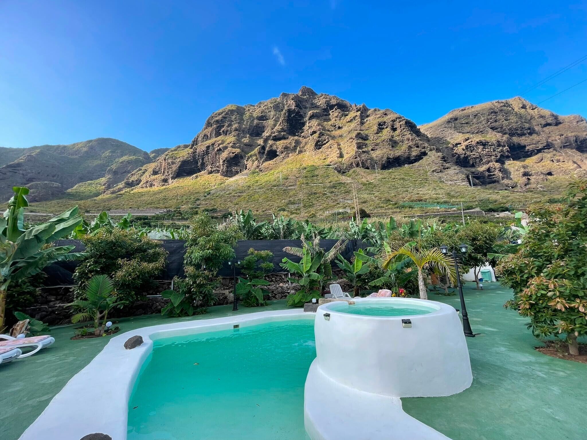 Una piscina amb les muntanyes al fons, envoltada de vegetació exuberant i un cel blau.