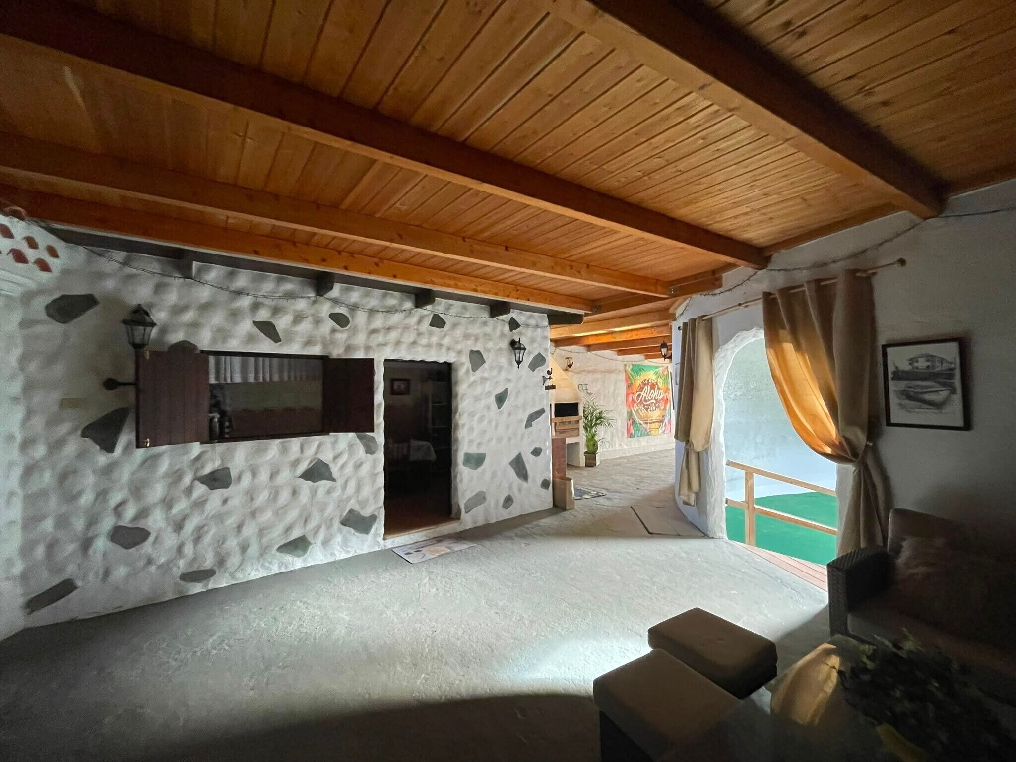 A room with a stone-patterned wall, a wooden ceiling, a window with curtains, and a small seating area.