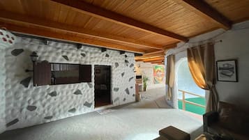 A room with a stone-patterned wall, a wooden ceiling, a window with curtains, and a small seating area.