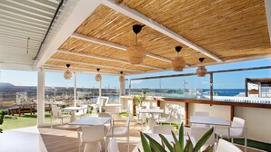 Un espacio de comedor al aire libre con mobiliario blanco, techo de madera y vistas al océano.