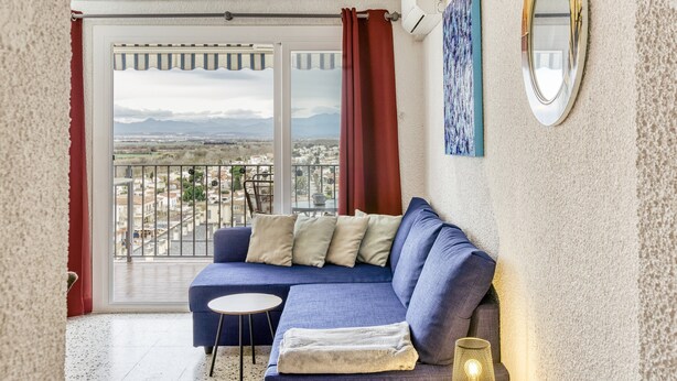 Un balcón con vistas a un paisaje urbano y montañas, un sofá azul con cojines, una pequeña mesa redonda y un espejo fijado a la pared.