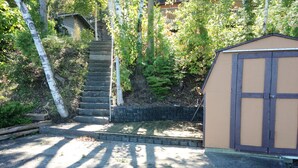 A covered outdoor area with a wooden deck, a small shed, and a staircase leading up to a house.
