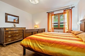A bedroom with a wooden dresser, a bed with striped bedding, a window with curtains, and a mirror on the wall. - Apartment 'Collini Le Betulle' with Mountain View, Private Garden and Balcony (Spiazzo)