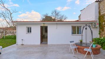 A small, single-story building with a tiled roof, a door, a window, and a small patio area with a table and chairs.