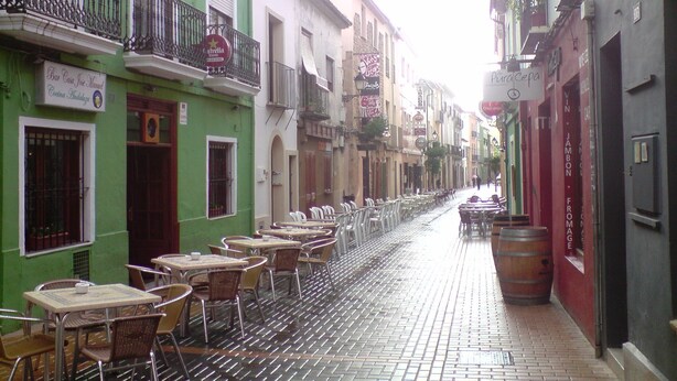 Una calle estrecha con mesas al aire libre, un edificio verde con letrero de bar y un edificio rojo con un barril.