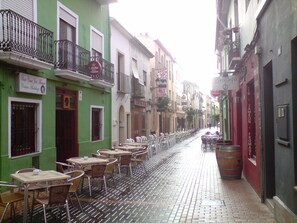 A narrow street with outdoor seating, a green building with a sign for a bar, and a red building with a barrel.
