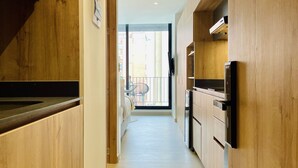 Un couloir étroit aux murs en bois, un coin cuisine avec des placards et un réfrigérateur, et un balcon avec une chaise et une table.