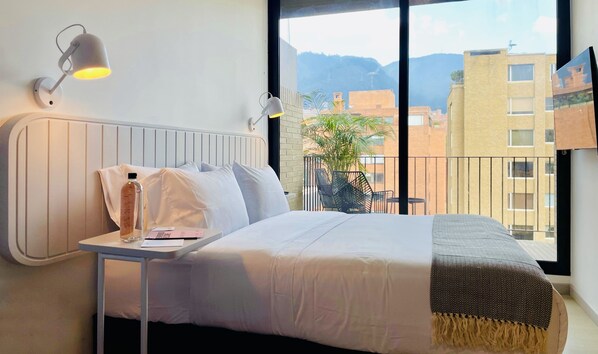 Habitación de hotel con cama, mesita de noche, lámpara y vistas a la ciudad y las montañas.