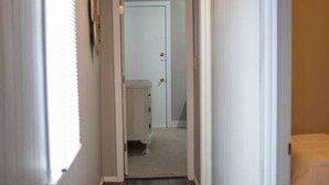 A narrow hallway with wooden flooring, white walls, and a door at the end leading to a bedroom with a bed. - Charming Magnolia Apartment (Magnolia)