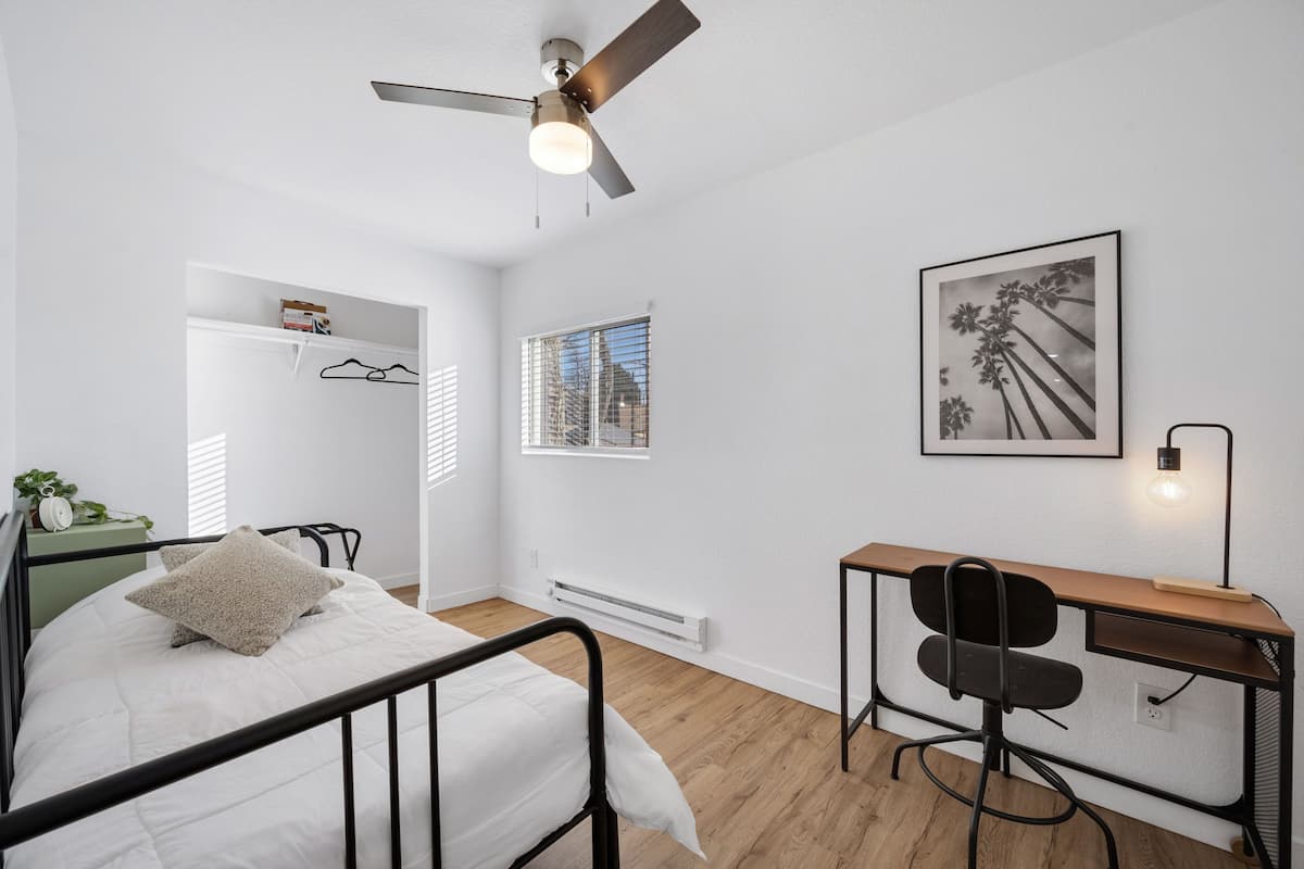 Een slaapkamer met een bed, een bureau, een stoel, een plafondventilator, een radiator, een raam en een ingelijst kunstwerk aan de muur.