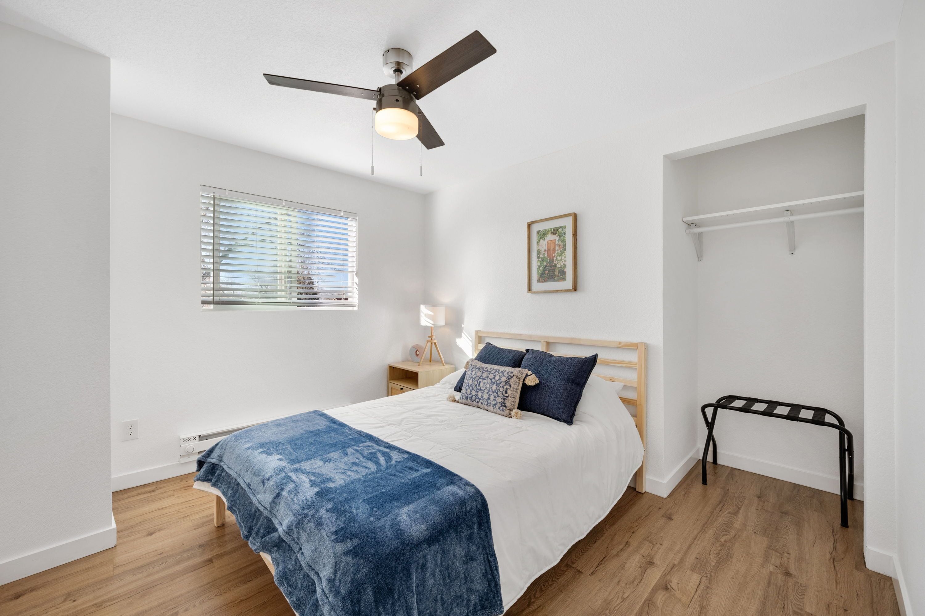 A bedroom with a bed, a ceiling fan, a window with blinds, a bedside table with a lamp, and a wall-mounted shelf.