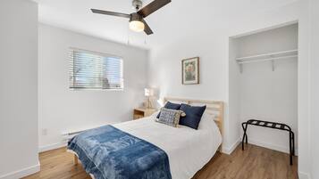 A bedroom with a bed, a ceiling fan, a window with blinds, a bedside table with a lamp, and a wall-mounted shelf.