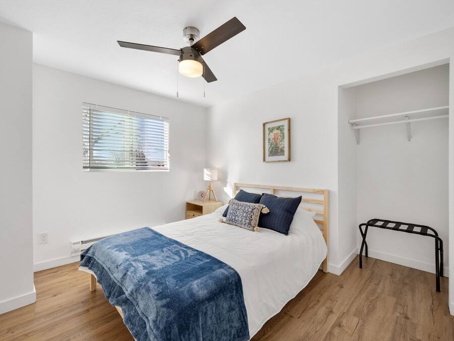 A bedroom with a bed, a ceiling fan, a window with blinds, a bedside table with a lamp, and a wall-mounted shelf.