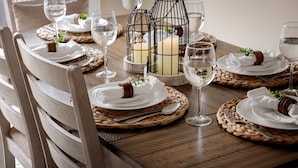Une table de salle à manger en bois, dressée avec de l’argenterie, de la vaisselle blanche et des verres, entourée de sets de table en fibres tressées et de bougeoirs.