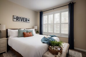 A bedroom with a large bed, a bedside table with a lamp, a window with curtains, and a wall sign that says "RIVER".