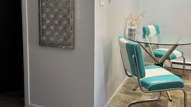 Un coin repas avec une table en verre et des chaises de couleur turquoise, un panneau métallique décoratif sur le mur et un tableau au-dessus de la table.
