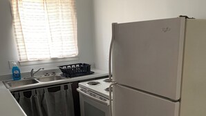 Une cuisine avec un réfrigérateur, une cuisinière et un évier blancs. Il y a une fenêtre avec des stores et un ventilateur de plafond.
